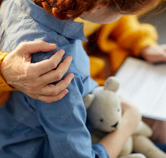 une main adulte se pose sur l'epaule d'un enfant de dos qui tient son doudou