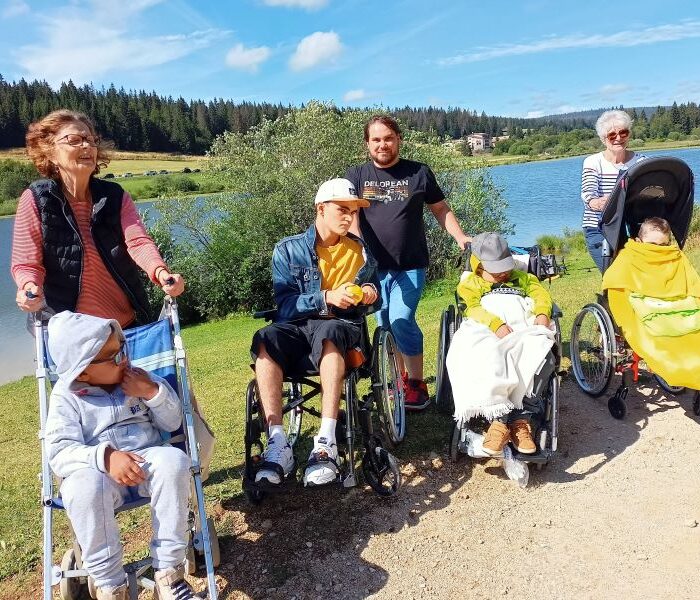 Grup de 4 enfanst en fauteuil avec 3 benevoles en balade sur un chemin avec un lac à l'arriere