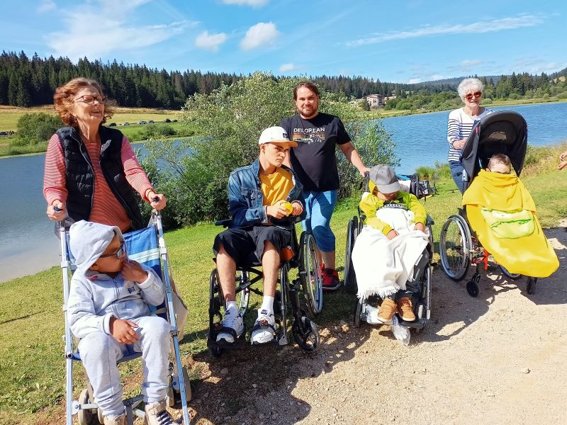 Grup de 4 enfanst en fauteuil avec 3 benevoles en balade sur un chemin avec un lac à l'arriere